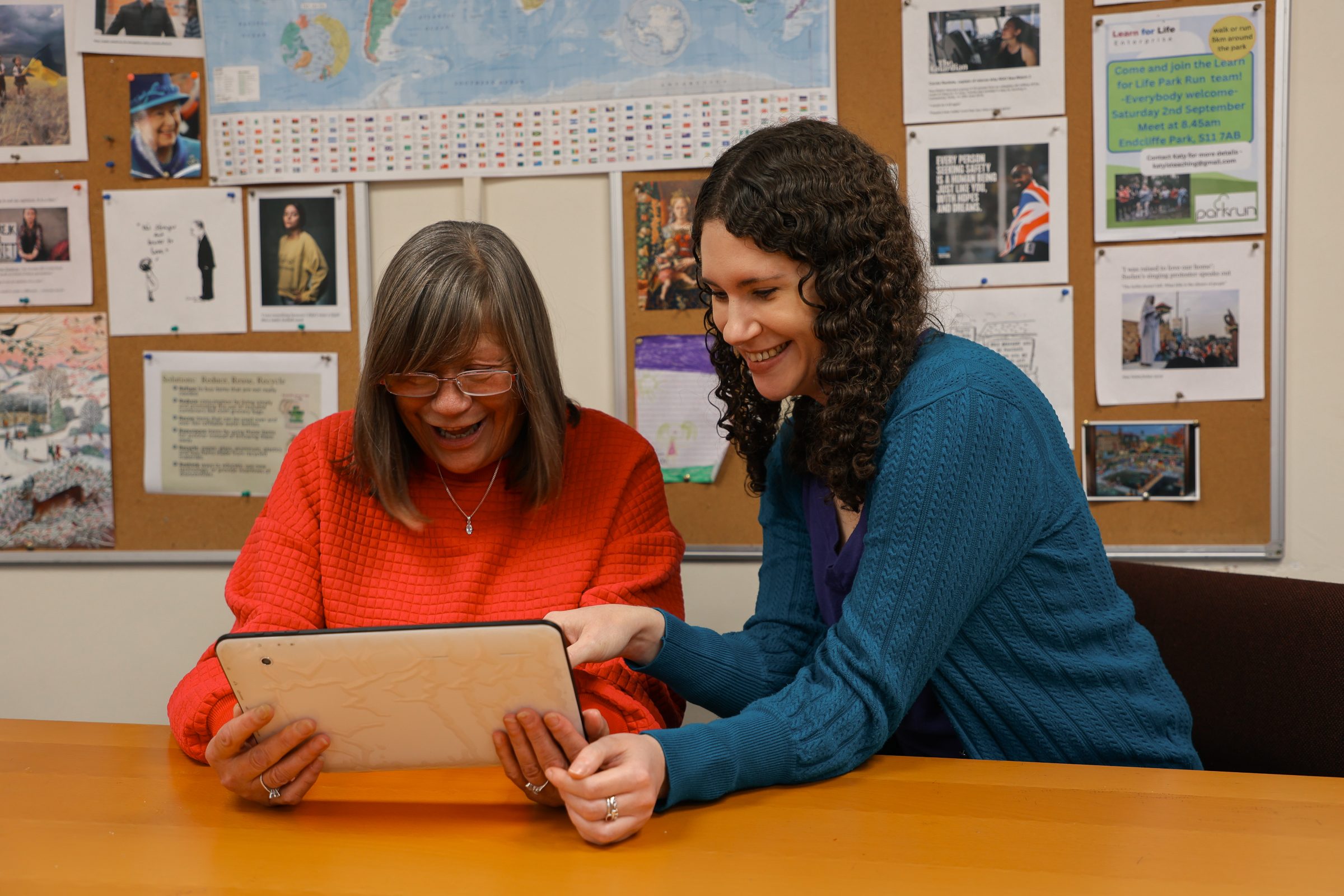 Two women smiling while looking at a tablet in an office.