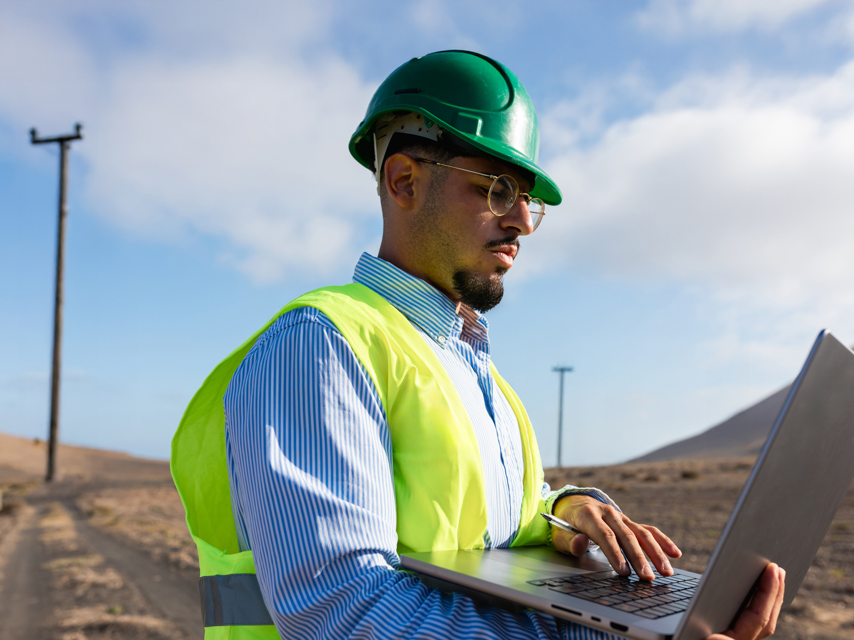 Engineer in a hard hat and hi-vis vest using a laptop outdoors.